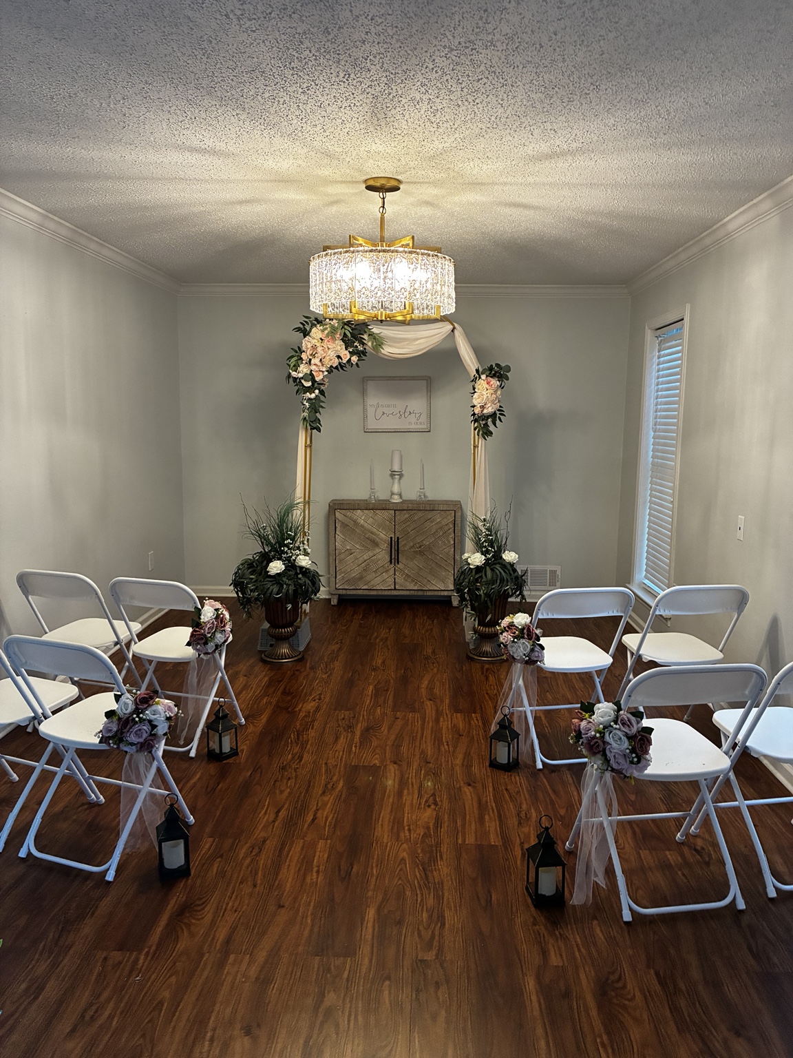 The ceremony room set for a wedding, with a floral arch under a crystal chandelier, white chairs, and lanterns along the aisle